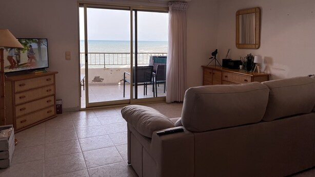 Ein Wohnzimmer mit einer Couch, einem Fernseher, einer Holzkommode und einem Balkon mit Blick auf den Strand.