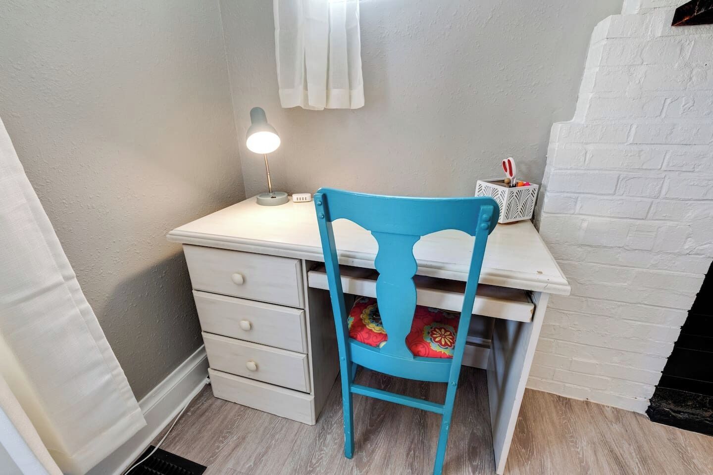 Et lille, hvidt skrivebord med en turkis stol, en skrivebordslampe og en dekorativ æske.