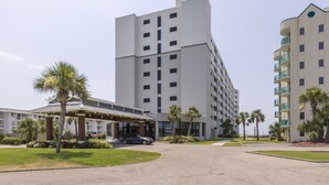 Un hotel de varios pisos con entrada central, palmeras y un área de estacionamiento techada.