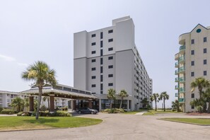 Een hotel met meerdere verdiepingen, een centraal gelegen ingang, palmbomen en een overdekte parkeerplaats.