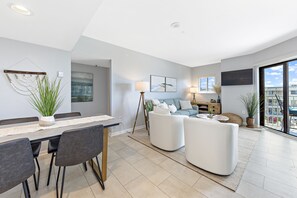 A modern living room with a dining area, a sofa set, and a large window offering a view of the city. - Refresh, Relax, and Recharge!  Family-sized Resort Beach condo. (Gulf Shores)
