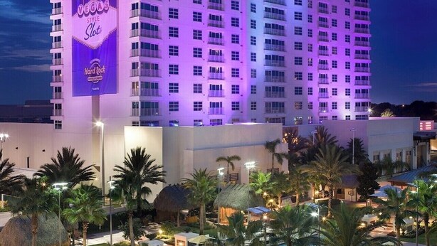Um grande hotel com uma placa chamativa do Cassino Hard Rock, área de piscina com espreguiçadeiras e palmeiras.