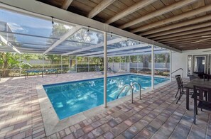 Una piscina exterior techada con un patio de ladrillo, una mesa y sillas, y vistas de la vegetación circundante.
