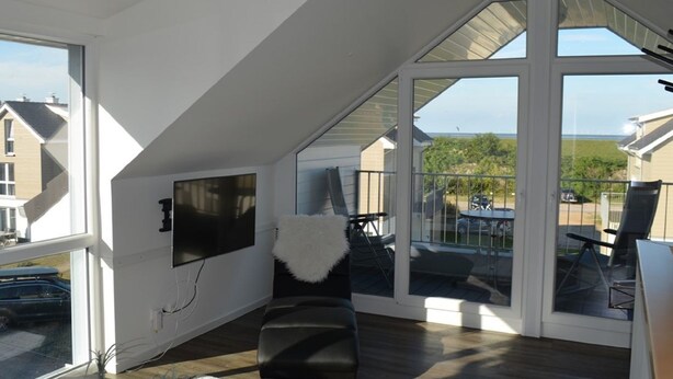 A modern room with large windows, a flat-screen TV, and a view of a residential area. - Haus Nr. 37, Ferienhaus Deichgraf - Gepflegtes Ferienhaus, Sauna, Süd-terrasse, Loggia mit Meerblick (Wangerland)