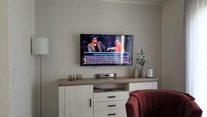 A modern living room with a flat-screen TV mounted on the wall, a white cabinet with drawers, a red armchair, and a floor lamp. - Haus Nr. 37, Ferienhaus Deichgraf - Gepflegtes Ferienhaus, Sauna, Süd-terrasse, Loggia mit Meerblick (Wangerland)