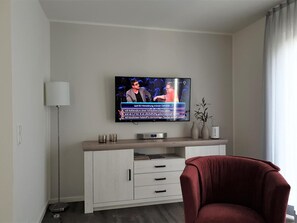 Ein modernes Wohnzimmer mit einem flachbildfernseher an der Wand, einem weißen Schrank mit Schubladen, einem roten Sessel und einer Stehlampe.