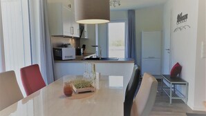 A modern kitchen with a built-in oven, a dining area with a glass table, and a red chair. - Haus Nr. 37, Ferienhaus Deichgraf - Gepflegtes Ferienhaus, Sauna, Süd-terrasse, Loggia mit Meerblick (Wangerland)
