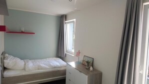 A bedroom with a bed, a dresser, a window with curtains, and a red shelf with a glass and a picture frame. - Haus Nr. 37, Ferienhaus Deichgraf - Gepflegtes Ferienhaus, Sauna, Süd-terrasse, Loggia mit Meerblick (Wangerland)