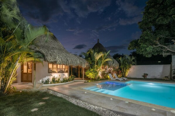 A thatched-roof house with a swimming pool, surrounded by palm trees and a well-maintained lawn.