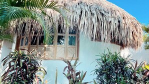Una casa con techo de paja, ventana de madera y plantas tropicales alrededor.