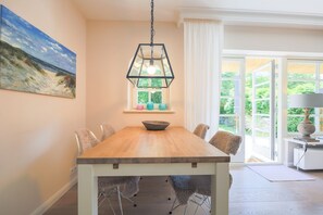 A dining area with a wooden table, chairs, a hanging light fixture, a painting, and a window with curtains. - Reethüs, Deutschland (Sylt)