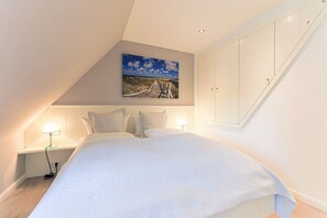 A bedroom with a large bed, a nightstand with a lamp, a wardrobe, and a painting of a landscape on the wall. - Reethüs, Deutschland (Sylt)