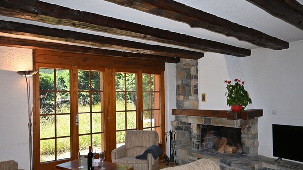 Ein gemütliches Wohnzimmer mit Kamin aus Stein, Holzbalken und einem großen Fenster mit Blick auf Grünflächen.