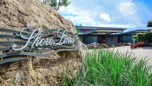 Une pancarte portant l’inscription « Shore Limes » est fixée à un rocher, entouré de verdure, avec un bâtiment moderne en arrière-plan.