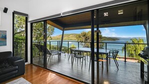 Un balcon spacieux avec une terrasse en bois, des sièges d’extérieur et une vue sur un plan d’eau et des collines.