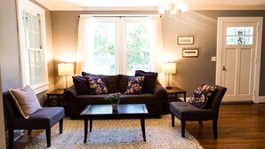 Un salon comprenant un canapé, deux fauteuils, une table basse et deux lampes. Il y a deux fenêtres avec des rideaux, un lustre et une porte avec des panneaux de verre.