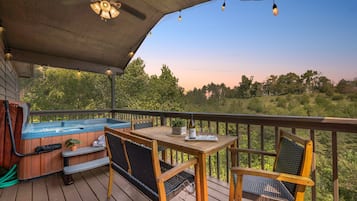 A wooden deck with a hot tub, a table, and chairs, overlooking a forested area.