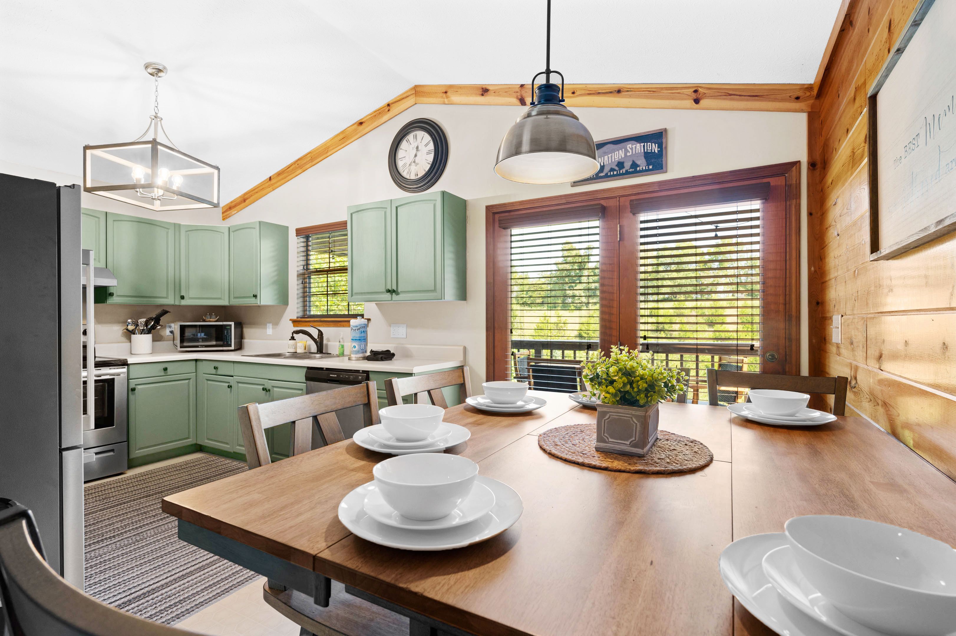 A kitchen with green cabinets, a wooden dining table set with white plates and bowls, a hanging light fixture, and a window with blinds.