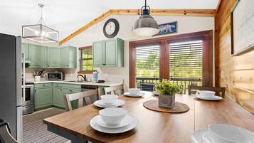 A kitchen with green cabinets, a wooden dining table set with white plates and bowls, a hanging light fixture, and a window with blinds.