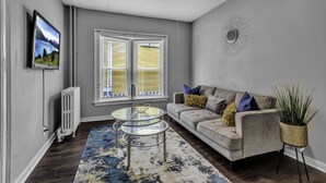 A living room with a gray sofa, a glass coffee table, a radiator, a window with yellow blinds, and a wall-mounted TV. - EXPLORE & RELAX Heart of the city,NEAR EVERYTHING! (Buffalo)