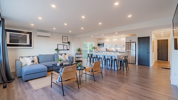 Un salon moderne avec un canapé, une table basse et des fauteuils, une cuisine avec des tabourets de bar et un coin repas avec une table et des chaises.
