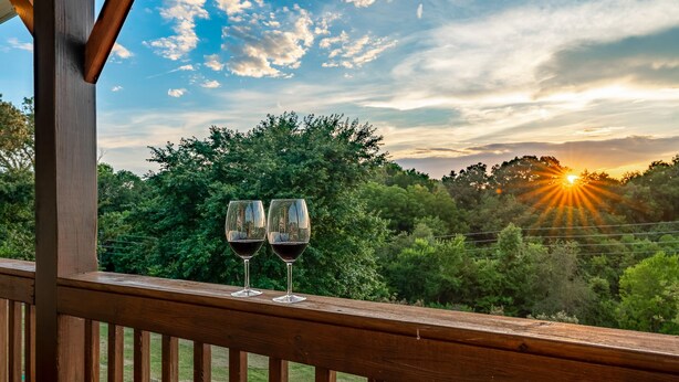 A wooden balcony with two wine glasses and a scenic view of a sunset over a forested landscape. - Private cabin with amenities ⭐️Location⭐️Spa Pool⭐️Hot tub⭐️Firepit⭐️Pets (Sevierville)