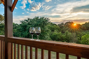 A wooden balcony with two wine glasses and a scenic view of a sunset over a forested landscape. - Private cabin with amenities ⭐️Location⭐️Spa Pool⭐️Hot tub⭐️Firepit⭐️Pets (Sevierville)
