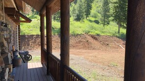 A wooden deck with a railing, a stone wall, and a view of a green landscape with trees and a dirt path. - Private Log Cabin with breathtaking views on 15 acres adjacent to forest service (Council)
