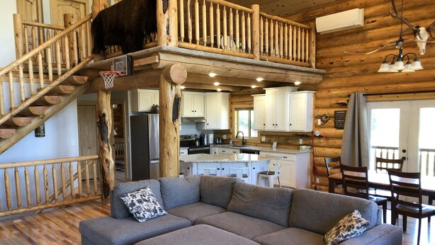 A log cabin interior with a gray sectional sofa, a wooden staircase, a kitchen area, and a dining area with chairs. - Private Log Cabin with breathtaking views on 15 acres adjacent to forest service (Council)
