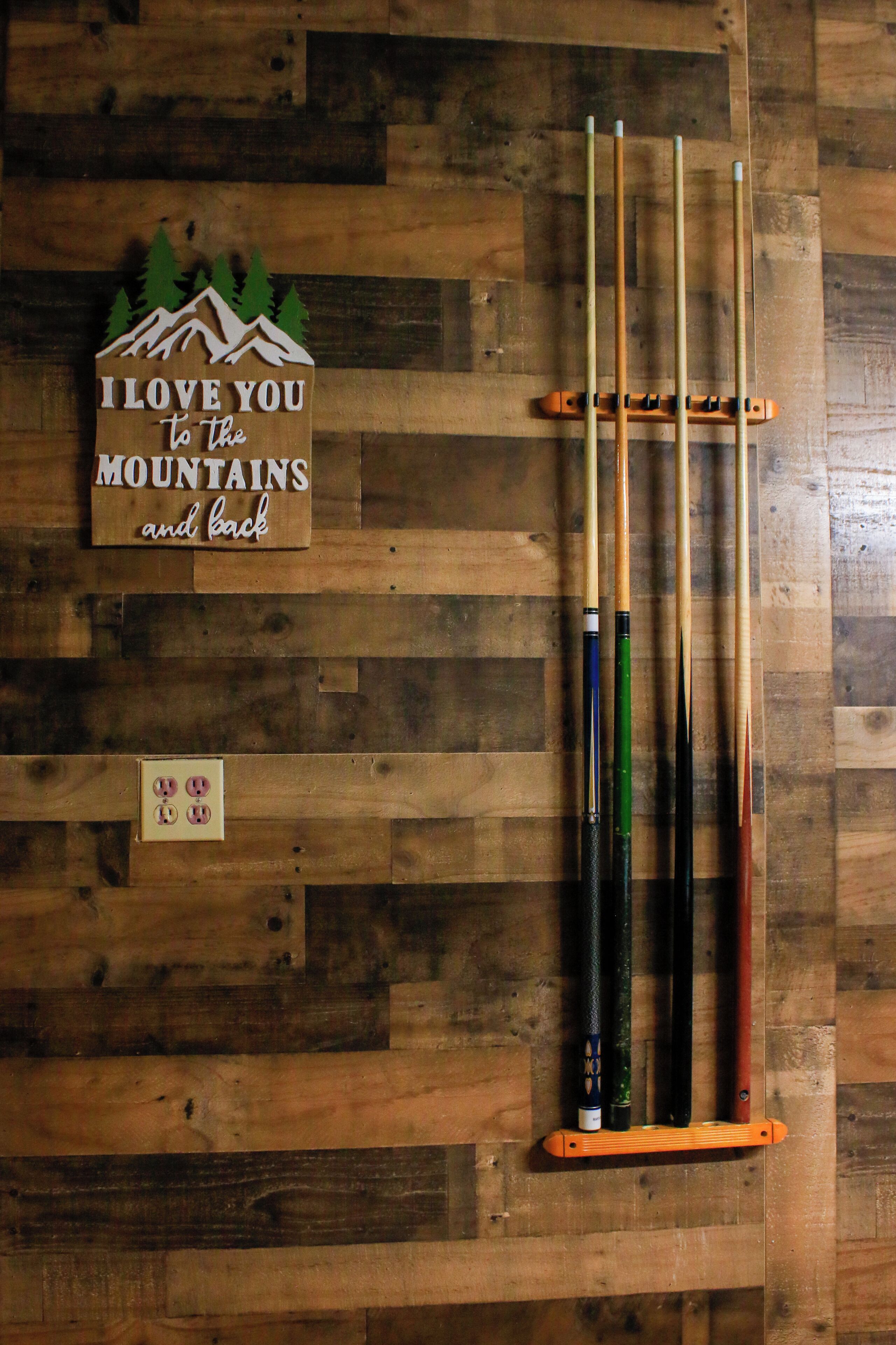 A wooden wall with a sign that reads "I love you to the mountains and back" and a pool cue rack with several cues.