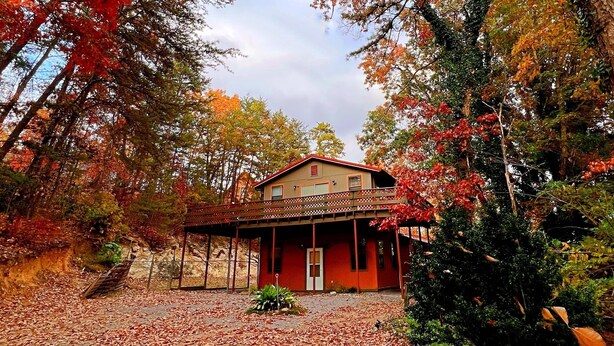 A house with a red exterior, a balcony, and a porch area surrounded by trees with autumn foliage.