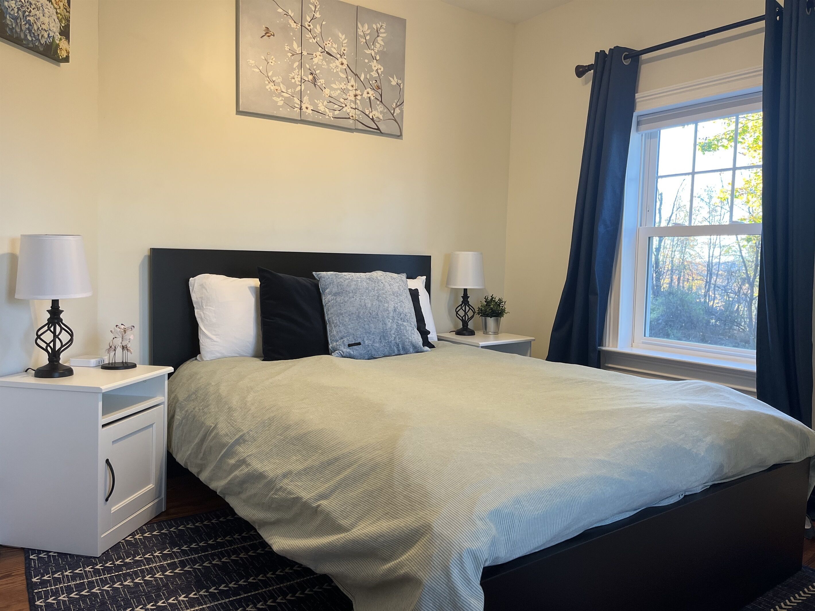 Een slaapkamer met een bed, twee nachtkastjes, een raam met gordijnen en een kunstwerk aan de muur.