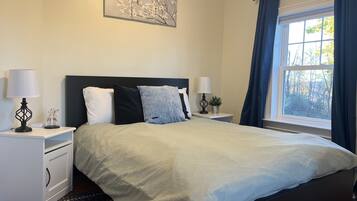 Een slaapkamer met een bed, twee nachtkastjes, een raam met gordijnen en een kunstwerk aan de muur.