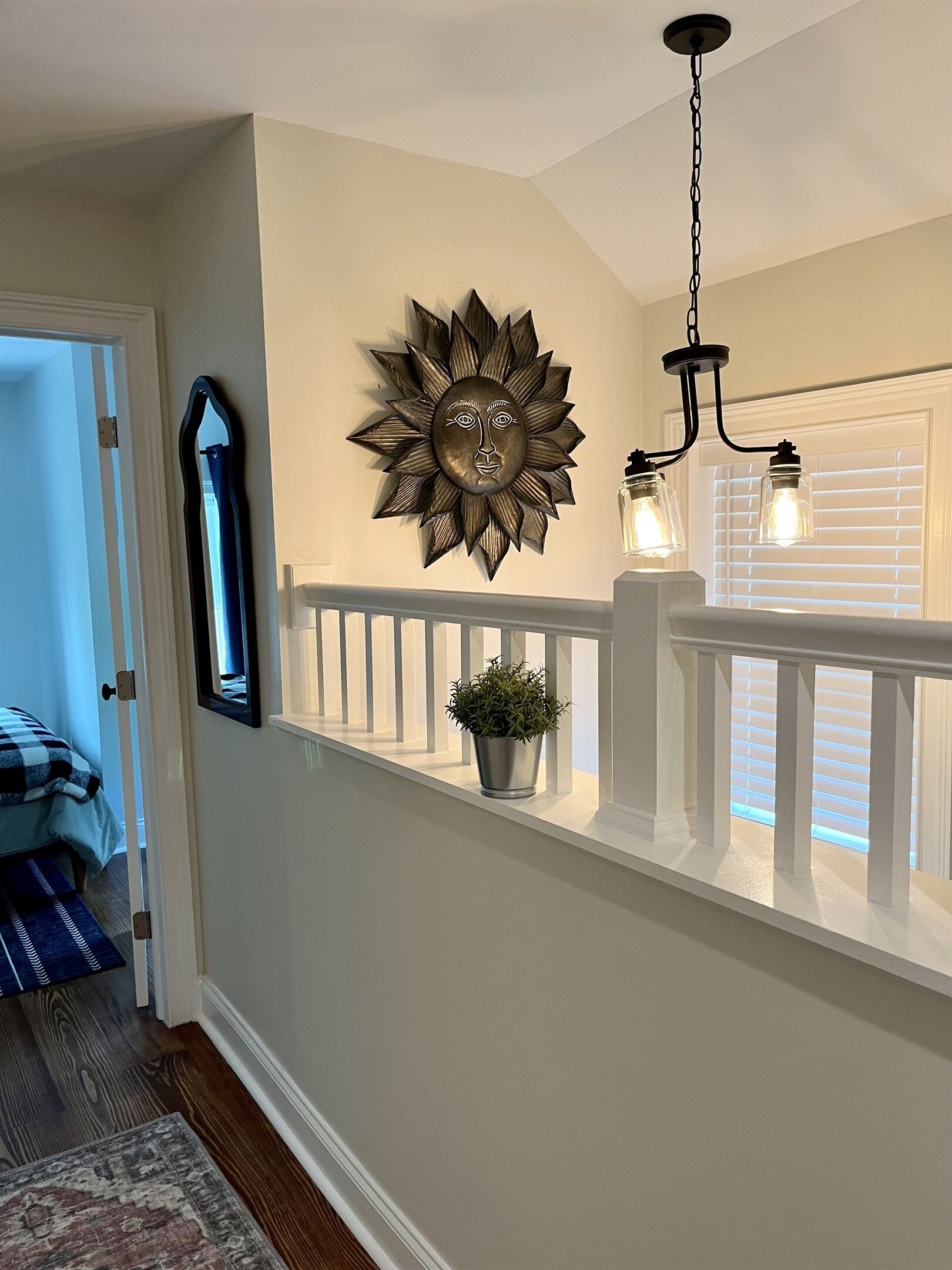 A hallway with a decorative sunburst wall art, a hanging light fixture, and a potted plant on a white railing.