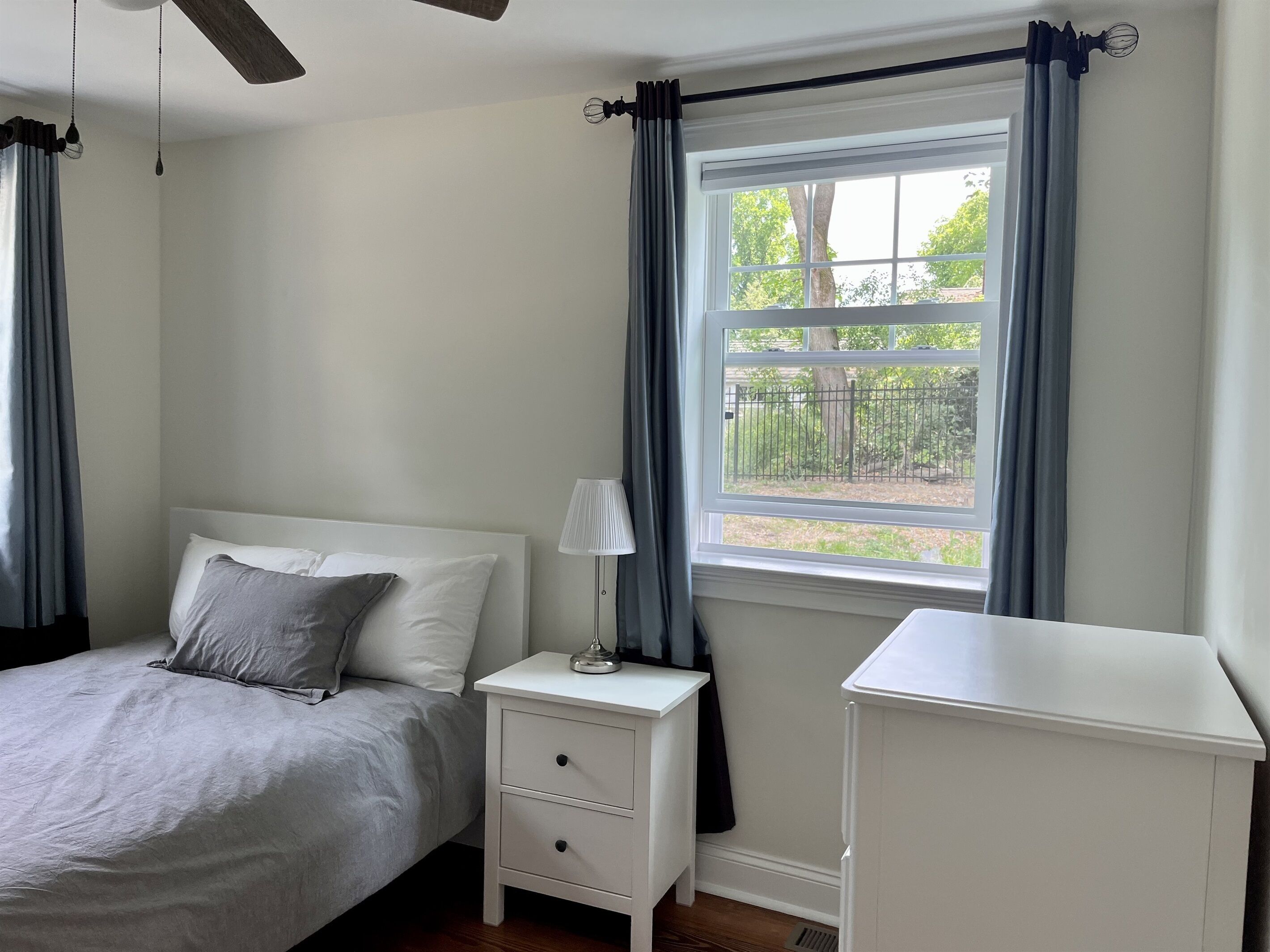 Ein Schlafzimmer mit einem Bett, einem Nachttisch, einer Lampe, einem Fenster mit Vorhängen und einem Deckenventilator.