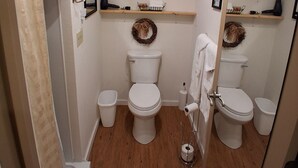 Une salle de bain avec deux toilettes, un rideau de douche, un miroir, une horloge et des étagères avec des serviettes.