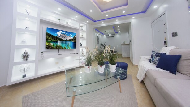 Una sala de estar moderna con una mesa de centro de vidrio, sofás blancos, un televisor de pantalla plana y plantas decorativas.