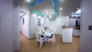 Un comedor moderno con una mesa y sillas blancas, una pequeña isla de cocina e iluminación empotrada en el techo.
