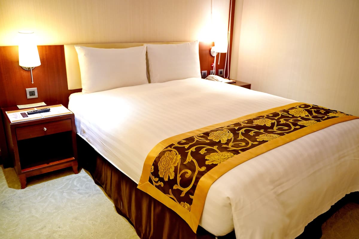 A neatly made bed with a white and gold bedspread, a bedside table with a lamp, and a telephone.