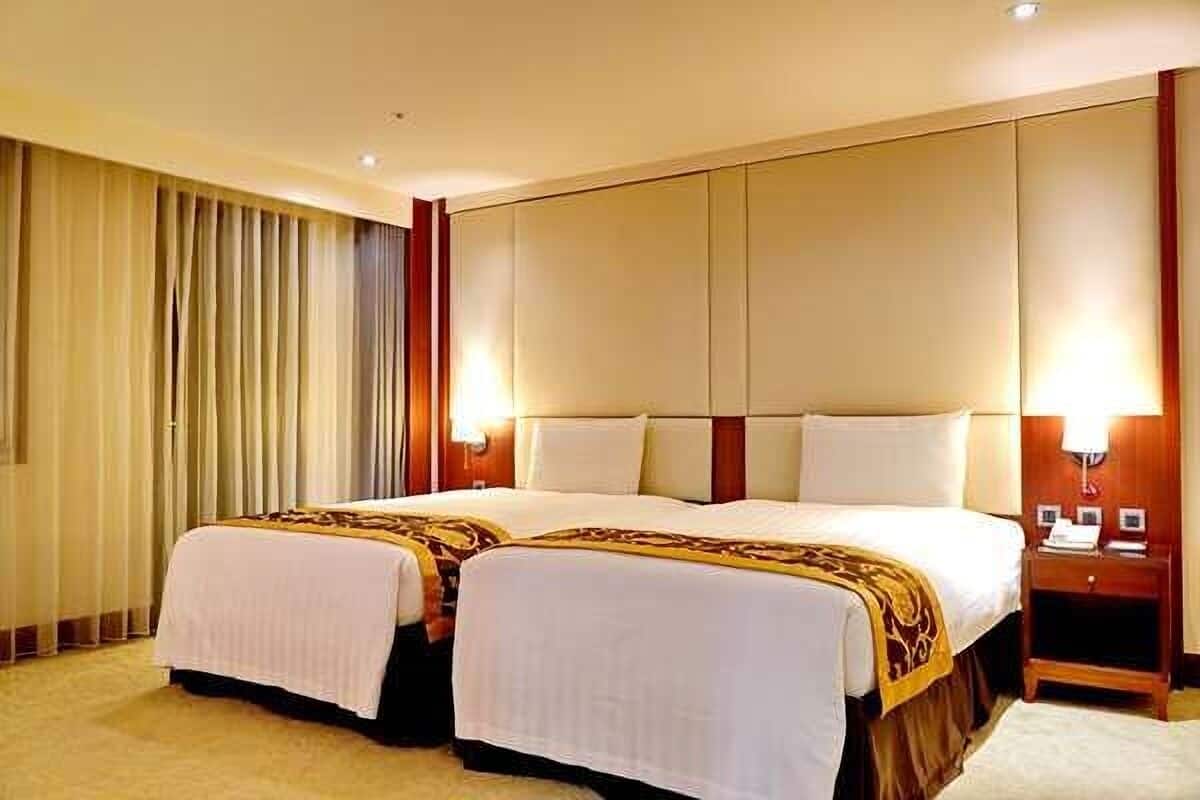 A hotel room with two beds, a large window with curtains, a bedside table with a lamp, and a nightstand with a phone and a book.