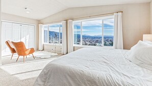 Un dormitorio con una cama grande, una silla y vistas a un paisaje urbano a través de amplios ventanales.