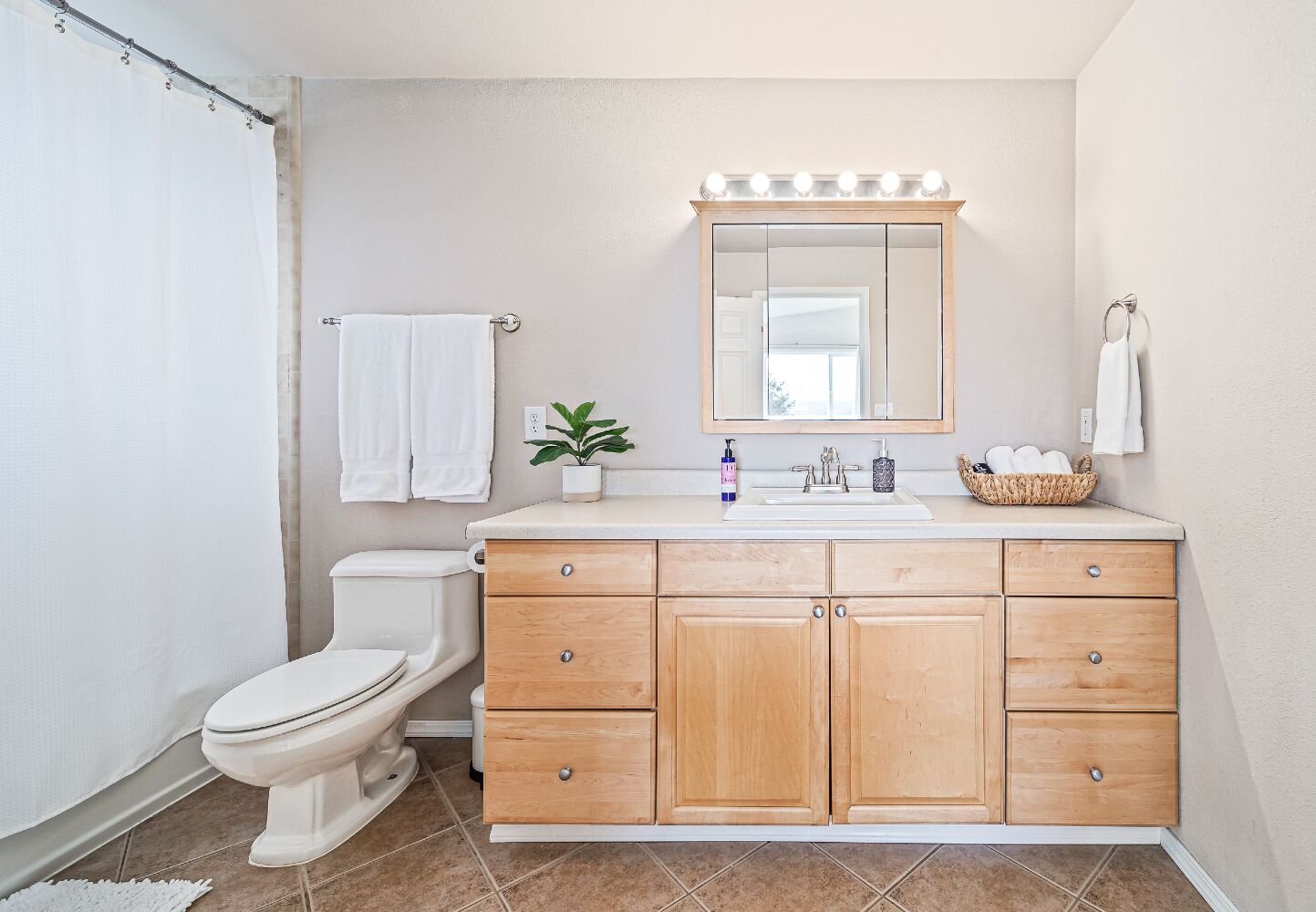 Une salle de bain avec des toilettes blanches, un meuble-lavabo en bois doté de tiroirs, un miroir avec éclairage, une petite plante et un panier rempli de serviettes.