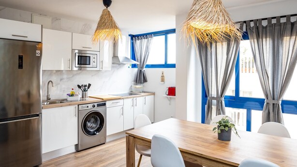 A modern kitchen with a stainless steel refrigerator, microwave, and washing machine. There is a wooden dining table with chairs and a hanging light fixture.
