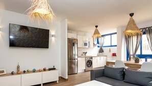 A modern living room with a grey sofa, white coffee table, and a TV mounted on the wall. There is a kitchen area with a washing machine and a dining table with chairs.
