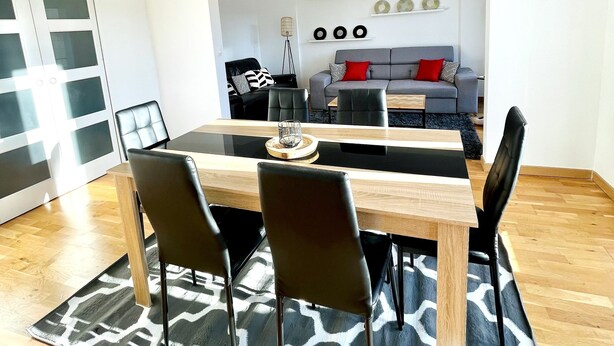 Uma zona de refeições moderna com uma mesa de madeira e cadeiras pretas, um tapete estampado e um sofá ao fundo.