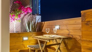 Un espacio de estar al aire libre con madera, una mesa y dos sillas, dos copas de vino y una maceta con flores rosas.