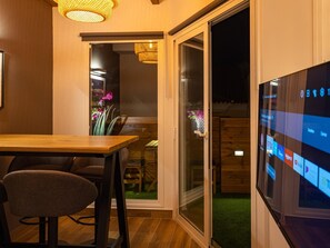 Een modern interieur met een houten eettafel, een comfortabele stoel, een televisie waarop verschillende zenders te zien zijn, en een schuifdeur naar een buitenruimte met een houten terras en een kleine tuin.