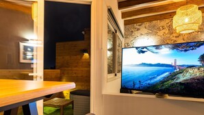 Una habitación con una mesa de comedor de madera, una silla cómoda, un televisor en la pared mostrando un paisaje y una cesta de mimbre en el suelo.