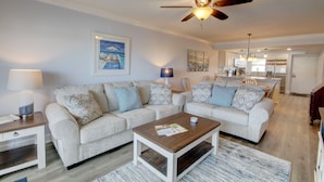 A living room with a sectional sofa, a coffee table, a ceiling fan, and a kitchen in the background. - El Matador 461 -  FREE TICKETS!! Your stay includes free tickets to many area attractions! (Fort Walton Beach)
