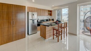 A modern kitchen with wooden cabinets, a stainless steel refrigerator, a built-in island with bar stools, and a large window offering a city view.